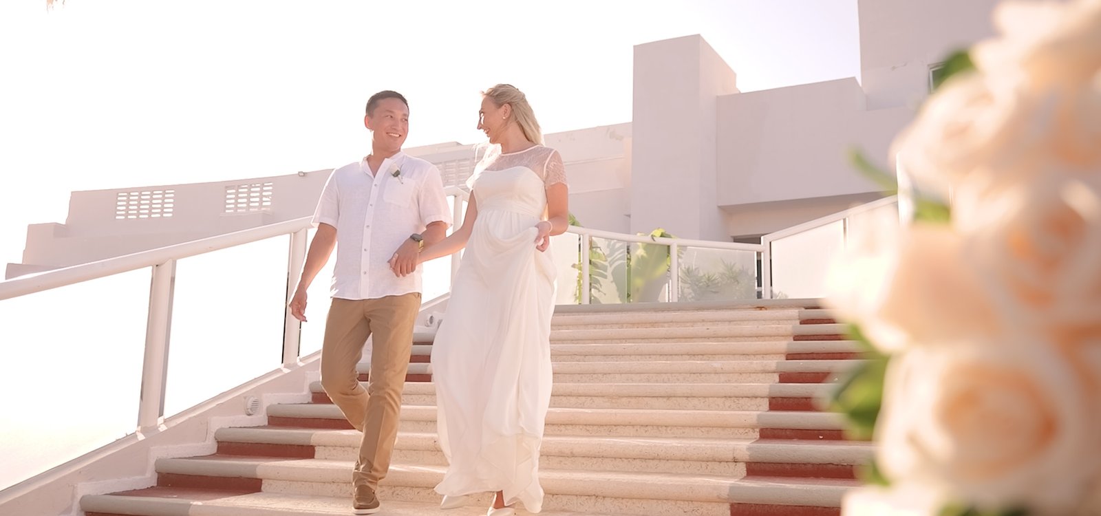 A couple of newlyweds smile under a pergola adorned with white flowers and warm lights. She is wearing a lace dress and he is in an elegant suit, both gazing at each other with love. Around them, a small group of guests is applauding and enjoying the moment in a cozy, nature-filled setting. The atmosphere is intimate and romantic, highlighting the closeness and happiness of the newlyweds.