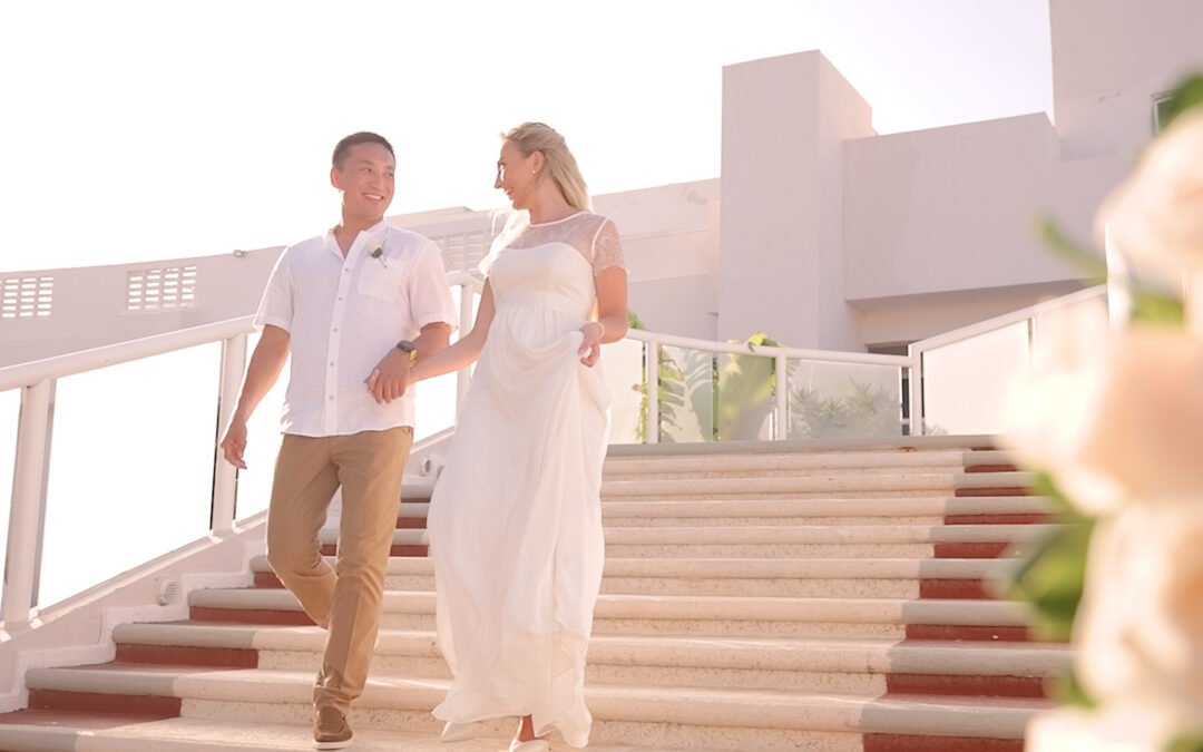 Una pareja de novios sonríe bajo una pérgola decorada con flores blancas y luces cálidas. Ella lleva un vestido de encaje y él un traje elegante, ambos mirándose con amor. Alrededor, se aprecia un pequeño grupo de invitados aplaudiendo y disfrutando del momento en un entorno acogedor y lleno de naturaleza. La atmósfera es íntima y romántica, destacando la cercanía y felicidad de los recién casados.