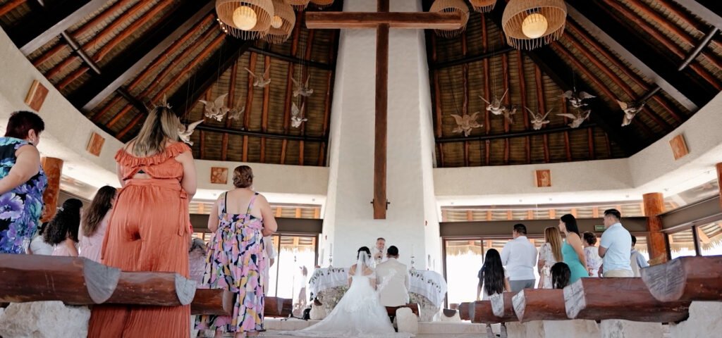 En la majestuosa Capilla del Hotel Xcaret, rodeada de exuberantes jardines y serenos cascadas, se lleva a cabo una boda católica con gracia y reverencia. Dentro de la capilla, adornada con velas y fragantes flores, la pareja se encuentra frente al altar, iluminada por el suave resplandor de vitrales que representan escenas de fe y amor. El sacerdote, con su voz resonante y tranquila, oficia la ceremonia mientras los invitados, vestidos con elegancia tropical, son testigos del intercambio de votos llenos de profunda devoción y compromiso. Afuera, el canto de los pájaros y el suave fluir de las aguas cercanas ofrecen un telón de fondo sereno, realzando la atmósfera sagrada de esta ocasión trascendental. Es un día donde la fe, el amor y la belleza natural se encuentran, marcando el inicio de un camino bendecido juntos en este impresionante paraíso de la Riviera Maya.