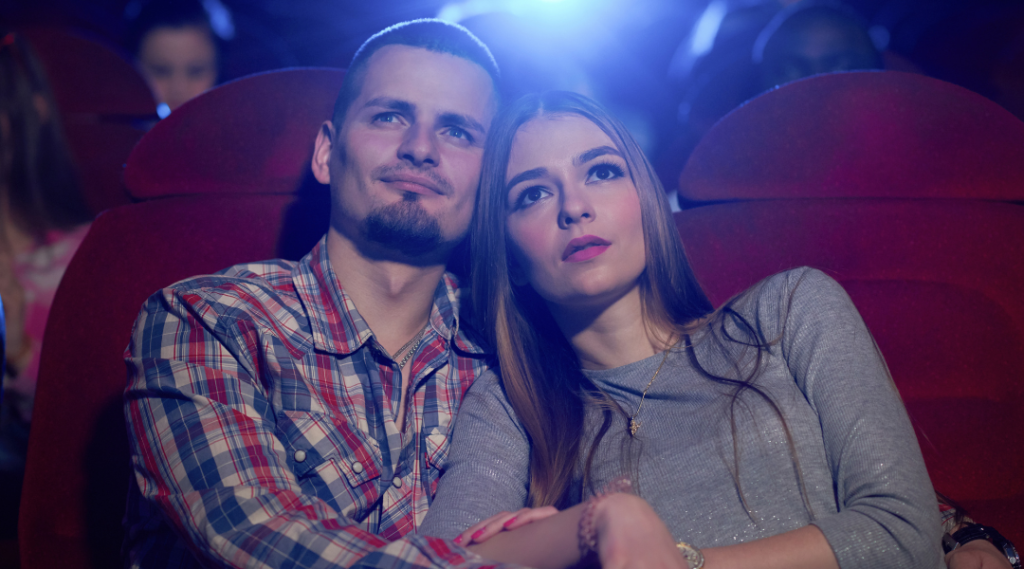 Una pareja sentada en un cine, ambos reclinados en sus asientos de felpa roja, compartiendo un balde de palomitas y una bebida. La sala está oscura, iluminada únicamente por la luz de la pantalla grande que proyecta una película emocionante. Los rostros de la pareja están iluminados por el resplandor de la pantalla, mostrando expresiones de asombro y alegría. Alrededor de ellos, otros espectadores están igualmente absortos en la película. El ambiente es de expectación y disfrute, con el suave murmullo de la audiencia y el sonido envolvente de la película llenando el espacio.