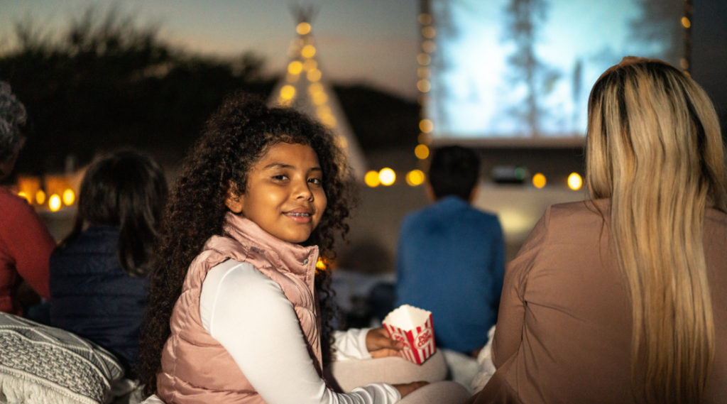 A couple sitting in a movie theater, both reclined in their plush red seats, sharing a bucket of popcorn and a drink. The room is dark, illuminated only by the light from the big screen projecting an exciting movie. The couple's faces are lit by the glow of the screen, showing expressions of amazement and joy. Around them, other viewers are equally absorbed in the film. The atmosphere is one of anticipation and enjoyment, with the soft murmur of the audience and the surround sound of the movie filling the space.