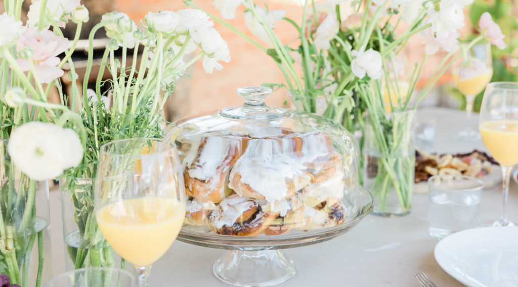 An elegantly decorated table for a wedding brunch in an outdoor setting. The table is covered with a white tablecloth and adorned with soft-colored floral arrangements. There is a variety of gourmet foods arranged, including fresh fruits, pastries, croissants, Eggs Benedict, a selection of cheeses and cured meats, and a standout dish of glazed cinnamon rolls. Champagne glasses and orange juice are ready for toasts. Guests, dressed in smart casual attire, are standing and sitting around the table, chatting and laughing while enjoying the food and company. In the background, a picturesque view of a garden or beach can be seen, completing the joyful and celebratory atmosphere of the event.