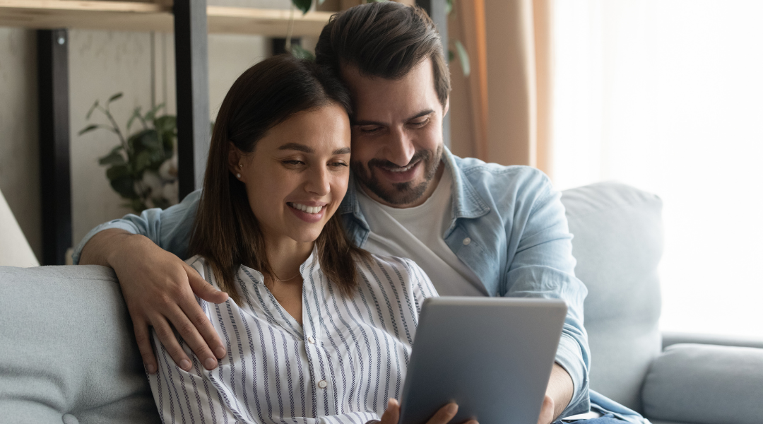 A couple sitting on a comfortable sofa in the living room, both smiling and laughing while watching a video on the television in front of them. The video shows happy moments from their wedding, with scenes of them exchanging vows, dancing, and celebrating with family and friends. The room is decorated in a cozy manner, with family photos on the walls and a coffee table with some magazines and a cup of tea. The soft light from a floor lamp adds a warm and nostalgic atmosphere to the scene.