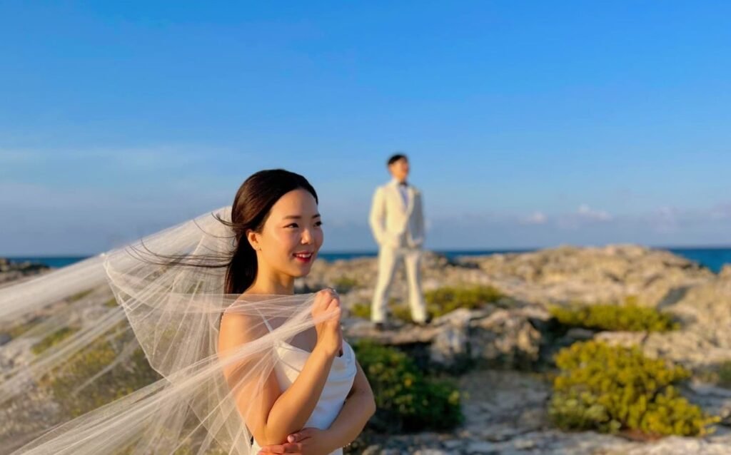 Couple posing on rocks by the sea; the bride smiles elegantly in the front, while the groom tenderly watches her from behind, creating a moment filled with love and intimacy.